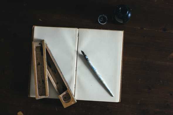 A calligraphy pen on top of a bright scetchbook on a very dark, wooden desk.